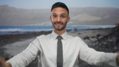 Gözlüklü ve sakallı genç adam kayalık bir plaj arka planında beyaz gömlek ve kravatla duruyor. Açık deniz ortamında kendine güvenen modern bir profesyoneli temsil ediyor.