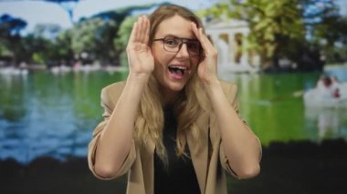 Neşeli bir şekilde gülümseyen bir kadın, güneşli yeşil bir parkta elleri ile yüzünü kapatarak, canlı bir arka plana karşı neşe sergiliyor..