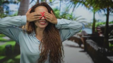 Uzun saçlı genç kadın iki eliyle gözlerini kapatıyor kırmızı tırnaklar ve yüzük gösteriyor, kollarını kaldırarak gülümsüyor; oyuncu bir sürpriz..