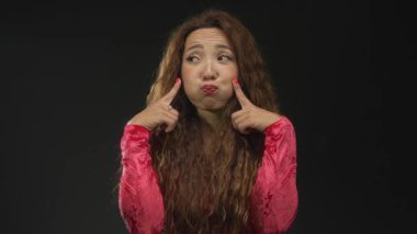 Stüdyoda koyu yeşil arka planda kırmızı kadife bluz giyerken işaret parmakları ve büzüşen dudaklarla yanaklara bastıran genç kadın;.