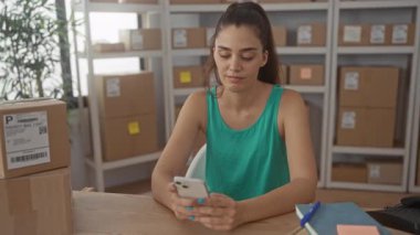 Kadın akıllı telefonu tutuyor ve bir binada istiflenmiş kahverengi paketler arasında çene dinleniyor; sessiz tefekkür.