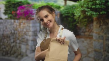 Önlüklü bir kadın gülümsüyor ve elinde bir kesekağıdıyla taş bir arkaplana yaslanıyor..