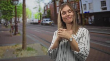 Kadın, binalar ve tramvay raylarıyla dolu hareketli şehir caddesinde dururken gülümseyip ellerini çırpıyor..