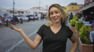 Genç sarışın kadın gülümseyen ve kamerayla meşgul olan bir kaldırım taşı sokak pazarında elini kaldırıyor; dostça karşılama.