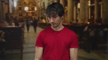 Genç adam boyalı camlarla aydınlatılmış bir kilise binasında ahşap sıraların yanında başını yana çeviriyor..