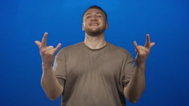 Gülümseyen genç adam, stüdyoda canlı mavi arkaplan karşısında hem eller hem de ön kollar ile kaya boynuzlarını gösteriyor; neşeli enerji.