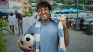 İspanyol adam futbol topu tutuyor ve atkı takıyor. Güneşli bir sokakta heyecanlı bir şekilde gülümsüyor..