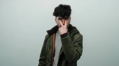 Siyah saçlı genç adam beyaz bir duvarın önünde tetikte bir jest yapıyor. Ceket giyiyor, tetikte ve odaklanmış olduğunu gösteren tuhaf bir ifade sergiliyor..