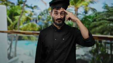 Siyah şef üniformalı bir adam arka planda palmiye ağaçları ve otelle dışarıda duruyor. Kafasını göstererek düşünceli bir ifade sergiliyor. Mutfak yaratıcılığını ima ediyor..
