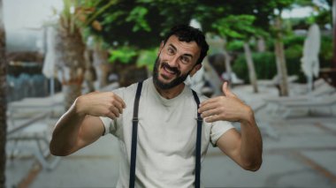 Sakallı İspanyol bir adam güneşli bir sokakta arka planda palmiye ağaçları olan beyaz bir gömlek ve pantolon askısı takarak coşkuyla el kol hareketi yapıyor..