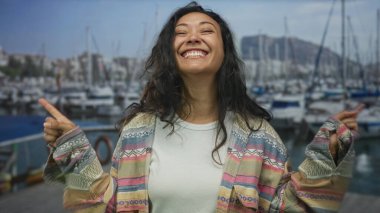 Woman smiling with arms crossed x and hands visible on a street pier by boats and a dock; carefree joy.