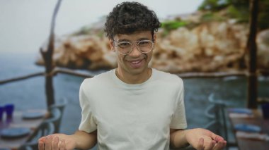 Yuvarlak gözlüklü ve beyaz gömlekli bir adam, açık denizde bir restoran terasında omuz silkme jestinde avuçlarını açık tutuyor; belirsizlik..