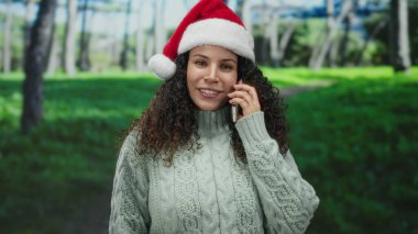 Noel Baba şapkası takan kadın telefonda konuşurken gülümsüyor yeşil kazak ve şenlik havasında..