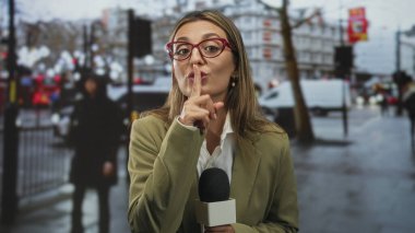 Kadın muhabir, sarışın, mikrofon tutan ve şehir sokaklarında sessizlik için parmaklarını dudaklarına bastıran; sakin otorite.