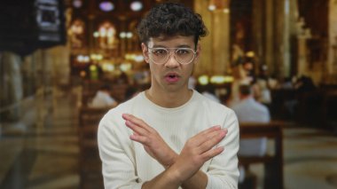 Çapraz kollu beyaz kazaklı bir adam kilisenin içinde vitray pencerelerin altında dikiliyor ve reddedildiğini gösteriyor..