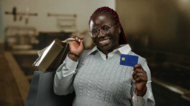 Mutfakta elinde alışveriş poşetleri ve kredi kartıyla gülümseyen, gözlük ve süveter takan bir kadın içeride başarılı bir alışveriş deneyimi sergiliyor..