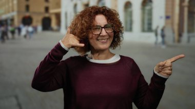 Elle işaret veren bir kadın, canlı bir şehir caddesinde duruyor, gülümsüyor ve gözlük takıyor..