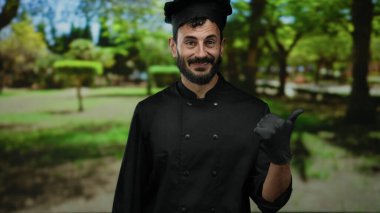Siyah üniformalı ve eldivenli İspanyol şef yeşil bir parkta ağaçlarla baş parmak işareti yapıyor, kameraya güvenle gülümsüyor..
