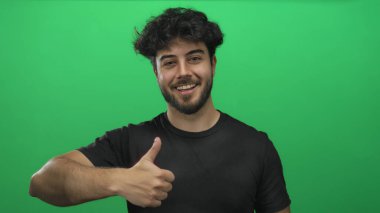 Sakallı gülümseyen genç bir adam neşeli ve kendinden emin bir ifadeyle olumlu ve olumlu bir görünüm sergileyen yeşil bir geçmişe başparmak sallıyor..