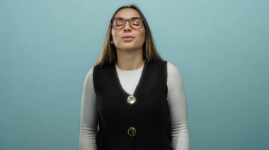 Gözlüklü ve yelekli kadın, izole edilmiş mavi arka plan duvarına karşı derin derin nefes alıyor, sakin ve odaklanmış, dingin bir atmosferde farkındalık ve rahatlamayı somutlaştırıyor..