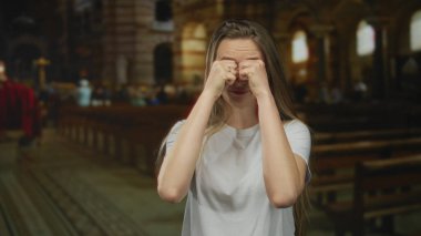Kilisedeki genç kadın uzun saçlı, sıradan bir kıyafetle, sıralarla çevrili kasvetli bir atmosfer yaratıyor..