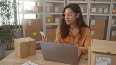 Kadın dizüstü bilgisayarda yazarken paket etiketlerini kontrol ediyor ve binadaki bir masada kutuları düzenliyor; küçük işletme odaklı.