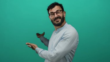 Sakallı, neşeli yeşil bir arka plana gülümseyen, el kol hareketi yapan, gözlüklü, açık mavi gömlekli, neşe saçan, parlak bir ortamda hoş geldin diyen bir adam..