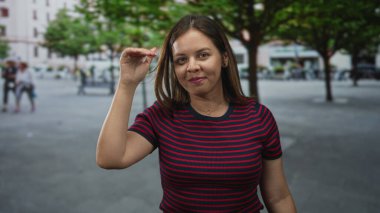 Beyaz kadın gülümseyen ve sağ elini sallayan ağaçlar ve meydanla görünür; tesadüfi bağ dostu.