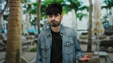 Sakallı, kot ceketli, terasta duran genç adam palmiye ağaçları arasında şüphesini dile getiriyor..