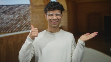 Gözlüklü adam, güneşli caddede binanın yanında gülümserken avuç içini işaret ediyor; kendine güven.