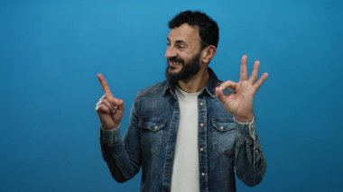 Sakallı, kot tişörtlü İspanyol bir adam. Tamam işareti yapıyor. Parlak mavi arka plana gülümsüyor..