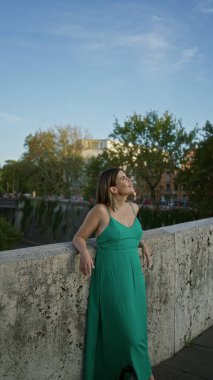 Roma 'da bir caddedeki taş köprü duvarına yaslanan yeşil elbiseli kadın, alacakaranlıkta nehrin kenarında bir sokak lambasının yanında, huzur içinde seyahat ediyor..