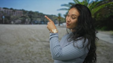 Kadın gülümserken ve kumlu sahilde binanın ve palmiye ağaçlarının yakınındaki kameraya bakarken kol saatini işaret ediyor; neşeli bir davet.