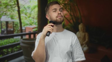 Adam bir otel binasında jileti çenesine tutarken sakalını elektrikli jiletle tıraş eder; kendine güvenini artırır.