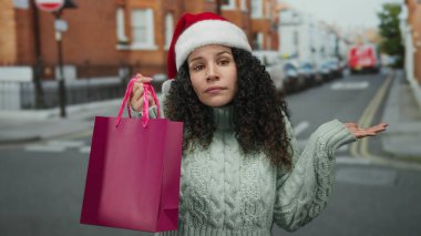 Noel Baba şapkası takan kadın, kış tatilinde şehir sokağında pembe alışveriş çantası tutarken şaşkın görünüyor..