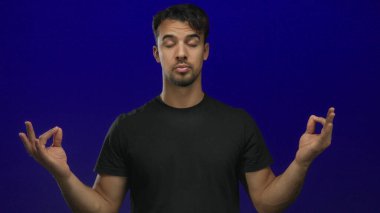 Genç İspanyol adam, sakin ve odaklanmış bir şekilde sakin bir arka plana karşı elleri ve gözleri kapalı meditasyon yapıyor..