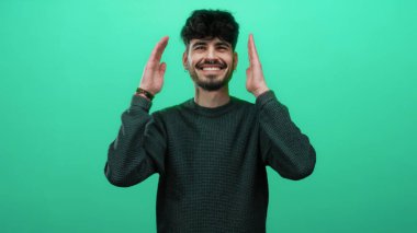 İzole edilmiş yeşil bir arka planda gülümseyen İspanyol genç adam, gündelik tarzını sergileyerek mola veriyor..