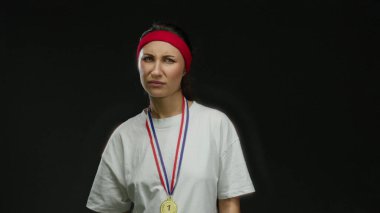 Madalya takan kadın, siyah arka plana karşı kaşlarını çatarak kırmızı saç bandı ve beyaz gömlekle kafa karışıklığını ifade ediyor, izole edilmiş stüdyo resmi, yüz ifadesini ve duygularını vurguluyor.