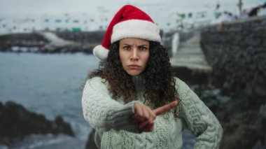 Noel Baba şapkası takan bir kadın, arka planda dalgalı deniz olan bir plajdaki kameraya kızgın bir şekilde bakıyor ve dışarıda keyifli bir hoşnutsuzluk taşıyor..
