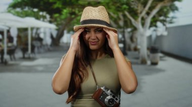 Güneşli bir günde başı ağrıyan bir kadın, şapka ve günlük giysiler giyiyor, turist kameraları asılı, arka planda ağaçlar ve bir kafe var..