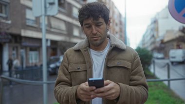 Latin bir adam şehir sokaklarında akıllı telefon kullanıyor, kahverengi bir ceket giyiyor, samimi bir anı yakalıyor ve yüzünde rahatlamış bir ifade var..