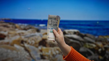 Sahilde elinde Peru banknotları tutan bir kadın. Arka planda mavi gökyüzü ve kayalık plajlarla finans ve seyahat sembolize ediyor..