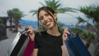 Kameraya gülümserken palmiye ağaçlarının altında kumsalda renkli alışveriş torbaları tutan kadın; eğlence heyecanı spontanelik.