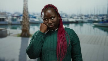 Pembe örgülü bir kadın arka planda tekneleri olan limanda yeşil bir kazak giyen, eli yüzü üzerinde düşünceli bir kadın..