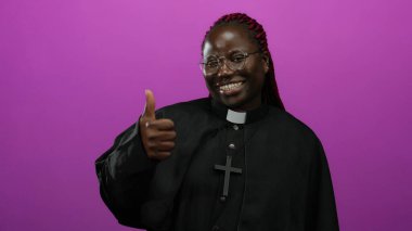 Kadın gülümsüyor ve parlak pembe arka plana karşı siyah rahip kıyafetiyle başparmak kaldırıyor, pozitiflik ve kendine güven yayıyor..