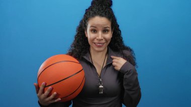 İspanyol kadın antrenör elinde bir basketbol topu tutarken boynunda bir ıslıkla canlı mavi bir arka plan tutuyor..