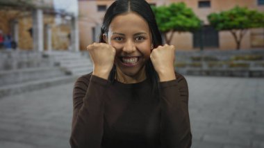 Kadın şehir sokak ortamında sevinçle kutlama yapıyor, arka planda ağaçlar ve binalarla dışarıda heyecan ve mutluluk ifade ediyor.