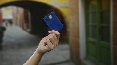 Elinde mavi bir kredi kartı olan beyaz bir adamın elinde yeşil kapılı ve arka planda kemerli bir caddede, şehir içi mali faaliyetlerini aktarıyor..