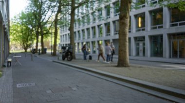 Bulanık insanlar, güneşli bir günde arka planda kadın ve erkeklerin yer aldığı modern binalarla, Avrupa 'nın ağaçlarla çevrili bir caddesinde yürüyorlar..