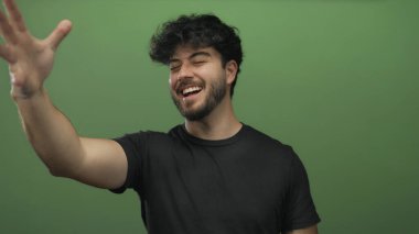 Sakallı genç İspanyol bir adam neşeli bir şekilde parmaklarıyla bir öpücük jesti gönderiyor. Canlı yeşil bir arka plan, neşeli bir ifade ve gündelik bir tarz sergiliyor..